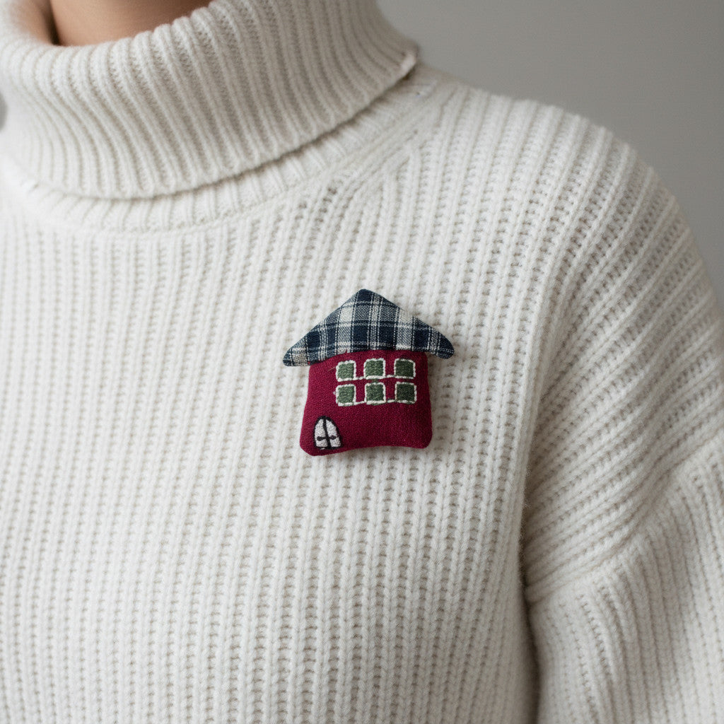 Magis Art Shop red textile house brooch with a plaid roof pinned on a white ribbed turtleneck sweater