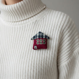 Magis Art Shop red textile house brooch with a plaid roof pinned on a white ribbed turtleneck sweater