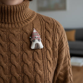 Magis Art Shop textile house brooch with a pink roof pinned to a brown cable-knit sweater